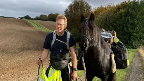 Roger Sewill during a trek in a field next to Scarlet. She is carrying his bags.