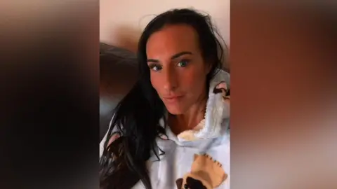 A selfie of Lisa Denton - she has long dark hair and stares into the camera. She is pictured sitting on a sofa, wearing a fluffy hoodie decorated with dogs.
