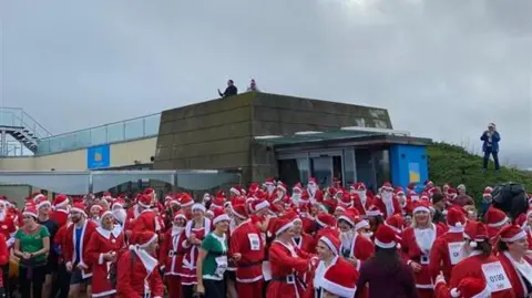 Linda Garnham A group of people dressed in red Santa suits and some people dressed as green elves. There is a building in the background and a grass bank on the right.