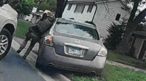 US District Court for the District of Minnesota Person in tactical gear stands beside a car on residential street, leaning toward driver’s side.