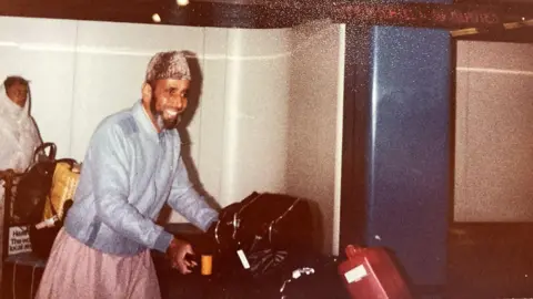BBC / Sabbiyah Pervez Mirza Khan walking through arrivals hall at London's Heathrow airport pushing his luggage on a trolley.