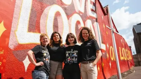 Khali Ackford Four women in black t shirts stand in front of a house painted with a red mural