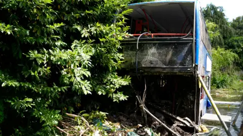 Blue double decker bus in river the water is just up to its wheels, the bus is badly damaged with windows smashed and debris strewn around the bus in the water.