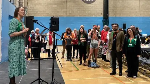 A Green Party member behind a microphone on a stage with others looking on