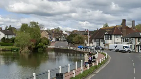 Google A village high street. A large pond sits on the side of a winding road.