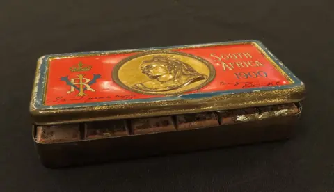 Lockdales Auctioneers and Valuers A tin box that is slightly open revealing an old chocolate bar inside. It is red on the top with a gold picture of Queen Victoria and the words "South Africa 1900".