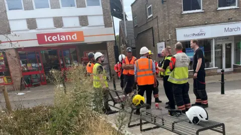 John Devine/BBC People wearing hard hats and high-vis jackets stand on a street opposite the Iceland shop, which appears with its red and white branding. There are two metal seating benches and dry-looking wild flowers planted in the foreground.