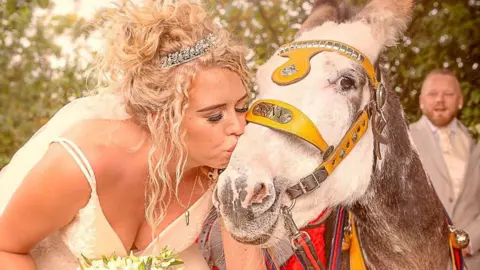 The Real Donkeys A bride in her wedding dress with her blond curly hair pinned up in a tiara kisses Sony the donkey.