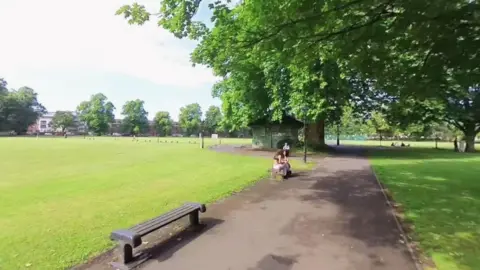 A view of Hoglands Park in Southampton on a sunny day. There are a few women sitting on a bench under big trees.