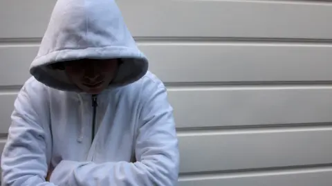 An unidentifiable teenage boy in a grey hoodie stands against a wall with his hood up and face in shadow. He has his arms crossed.