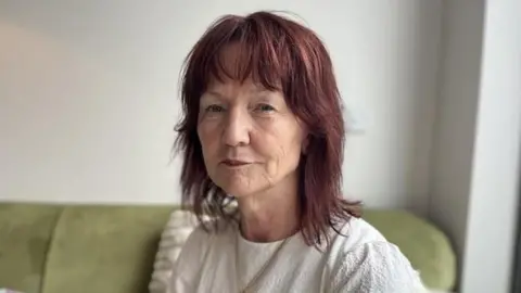 A woman with dark brown hair looking into the camera lens. She is wearing a white t-shirt and is sat on a green sofa with a white background.