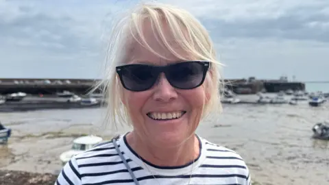 Elaine Davis pictured in Gorey Harbour. She is wearing a Breton-striped top, black sunglasses and has blonde hair. Behind her, the harbour is at low tide, and there are moored boats.
