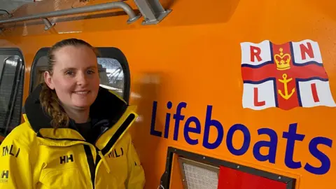 Chloe, a young woman with brown hair in a pony tail smiles wearing a yellow RNLI jacket and stands in front of an orange RNLI Lifeboat.