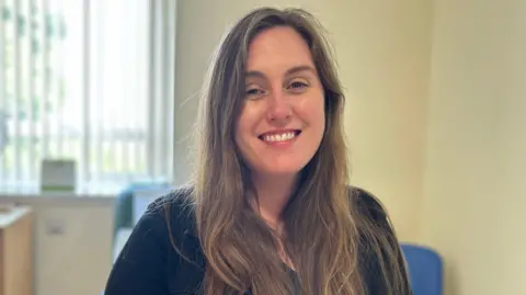 NHS Grampian A woman with long dark hair smiles into the camera. She is wearing a dark blue jumper and there is a window with blinds open behind her