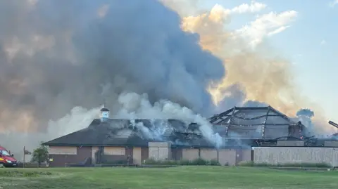 A golf club building with boarded up windows with smoke billowing upwards and part of the roof destroyed.