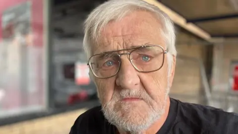 Pictured is Brian Dinnell. He has grey hair and a beard and wears aviator style glasses.