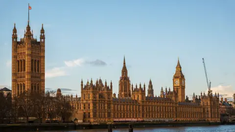Getty Images Houses of Parliament pictured during the day. The sky is light blue with only a couple of white clouds. The buildings are a gold/brown colour.