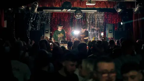 3flavourscornetto Dois DJs se apresentam atrás dos decks do palco. Ambos estão vestindo moletons escuros e bonés de beisebol. A pista de dança abaixo deles está lotada. Vários alto-falantes são colocados nas laterais do palco.