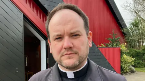 Shows a priest with a beard in clerical clothing in front of a red and black church