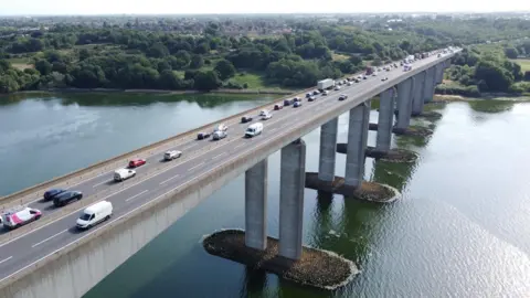 An aerial view of the Orwell Bridge showing the northern bank of the estuary. Traffic is queuing on the eastbound carriageway, where two lanes are open as vehicles approach the lane closure. Traffic is flowing on the westbound carriageway, where both lanes are also open. 