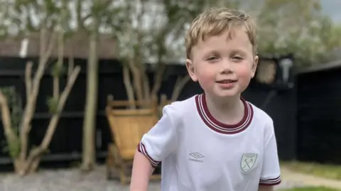 Lauren Carter/BBC Jesse standing in his garden holding the crossbar of a small football goal. He is wearing a white West Ham kit and is smiling.