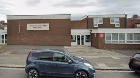 Google A brick building which has the sign of a cross on, with a sign reading: 'The Salvation Army Hartlepool'. Two cars are parked in front of the building on the road.