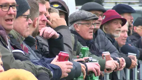 A line of men watch the match behind a metal barrier. A few are holding green Carlsberg cans, another another holds a Coca Cola can