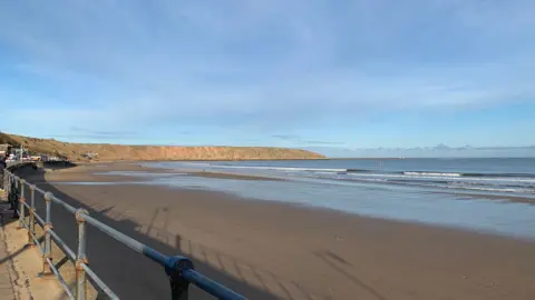 A wide coastal scene of Filey viewed from a promenade with a metal railing in the foreground. A broad sandy beach stretches out below, with shallow waves rolling in from a calm sea. Cliffs run along the far edge of the bay under a clear blue sky with light clouds.