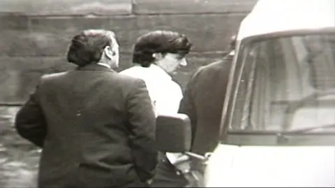 A black and white still from TV footage showing Peter Sullivan, who has thick black hair and who is wearing a white shirt, being led to a police van in handcuffs