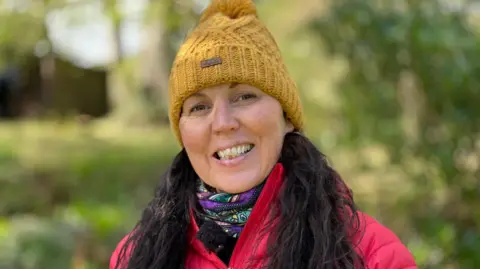 Kate Berquist is wearing a yellow woolly hat, and has long dark hair. She is wearing a red coat and is smiling, standing in a woodland surrounded by green grass and trees.