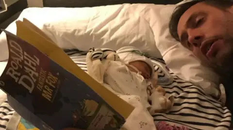 PA Media Ryan Lock, laying beside his baby daughter Ida in bed, reading a Roald Dahl's Fantastic Mr Fox.