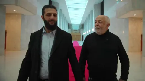 Ahmed al-Sharaa, wearing a black suit, walking with the BBC's Jeremy Bowen