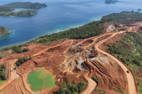 Global Witness A photograph taken in December 2024 shows mining activity on Kawei island, in Raja Ampat. On the island that is the main subject of the photograph, forest has been cleared to reveal brown earth, dirt roads built for mining vehicles and a pool where water from the mine collects.  