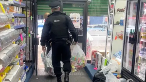 Blackburn and Darwen Council A police officer leaves a convenience stores carrying plastic see through bags that contain seized tobacco products. Inside the bags you can see the bright colours of vape boxes and in another bag the large cartons that contain cigarettes when bought in multipacks. 