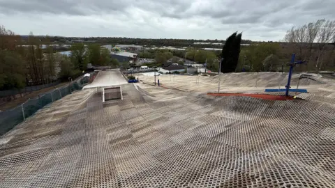 A dry ski slope, viewed from the top, with various mounds and obstacles for those using the facility. There are trees either side and there are buildings in the distnace.