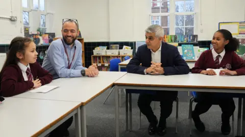 The mayor is sat at a school table in discussion with two female pupils wearing dark red cardigans, a white shirt and red striped tie. The headteacher is also at the table wearing a blue shirt and wearing glasses. They are smiling and talking. 