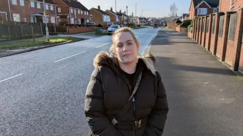 David Spereall/BBC A mum in her thirties with blonde hair. She is stood on a pavement next to a suburban road, wearing a brown coat.