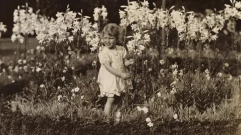 Royal Collection Enterprises Ltd 2025 | Royal Collection Trust A black and white photograph of a young girl in a summer dress in a meadow surrounded by flowers. 