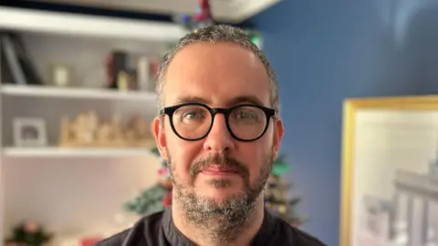 Keith Lawrence is a man with black and white hair and beard. He is wearing black glasses and a black grandad collar shirt. He is standing in his sitting room which is out of focus in the background. The walls are blue, there is a white cabinet, a white painting on the wall and a Christmas tree directly behind him.
