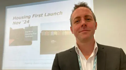 Robbie looks directly at the camera wearing a white shirt under a black suit jacket with a power point behind him saying "Launch of Housing First Nov '24"