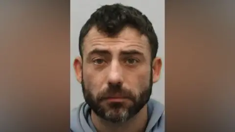 Mugshot of Rayne. He has short black hair and a thick black beard with white patches.