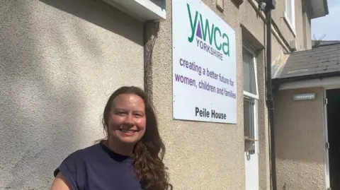 MICK LUNNEY/BBC A woman, Claire Harding, with long brown hair in a purple top stands outside a building. A sign on the wall behind her says it is Peile House, run by YWCA
