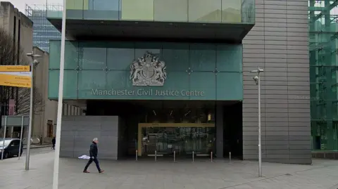 Entrance to Manchester Civil Justice Centre building with a man with grey hair walking past wearing a black coat, trousers and brown shoes.