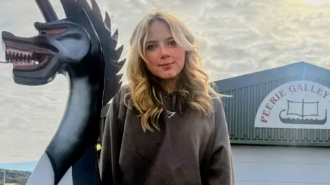 Erin is standing outside in front of shed and next to the dragon head of a mock Viking ship. She has long blonde hair and wearing a jumper. On the shed's corrugated metal wall is a sign. It reads: "Peerie Galley Shed".
