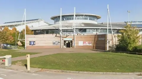 A large building has a round roof and is made from light orange bricks and is curved at the front. There is a grassy section to the front of the building.