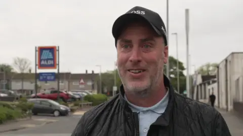 Billy Such smiling at the camera. To the left is an Aldi sign. 