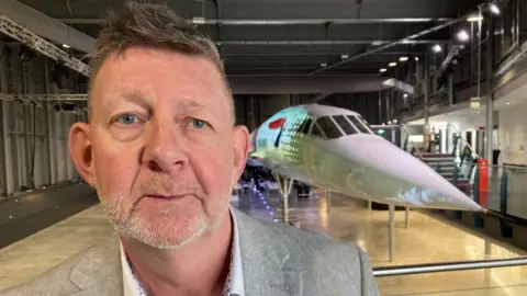 A man with quiffed brown hair and a short greying beard, wearing a greay suit jacket stands in front of the nose of a Concord jet on display at Aerospace Bristol
