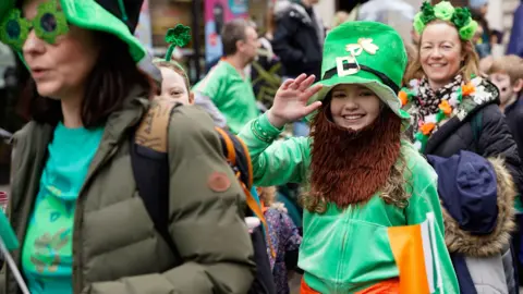 PA Media A young girl wearing a green leprechaun costume and ginger beard waves and smiles at the camera while other people wearing green flowers, shamrocks and garlands walk by