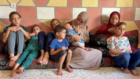 The Al-Najjar family sitting next to each other on floor cushions. The walls are painted in a pattern of different-coloured squares and the floor is stone tiles. The child on the left sits with their hands resting on their folded knees, next to them a boy is almost laying down, then another brother has his legs folded up. A smaller boy sits in front of them and is smiling to the side of the photo. Their mother sits in the middle wearing a brown headscarf and tunic, smiling at the baby she holds in her arms. Next to her is Malak, who is wearing a burgundy headscarf, with a smaller sibling in front of her with a patterned t-shirt.