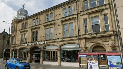 Google A Wetherspoons pub seen from the street. It is a three-storey sandstone building. A blue car is passing by. On the right is a bus stop. 